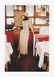 Woman in a beige cropped top, matching maxi skirt, and hat standing in an elegant restaurant interior with dining tables.