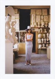 Model wearing a light blue ruffled outfit and structured wide-brim hat standing among plaster busts in a sculptor studio.