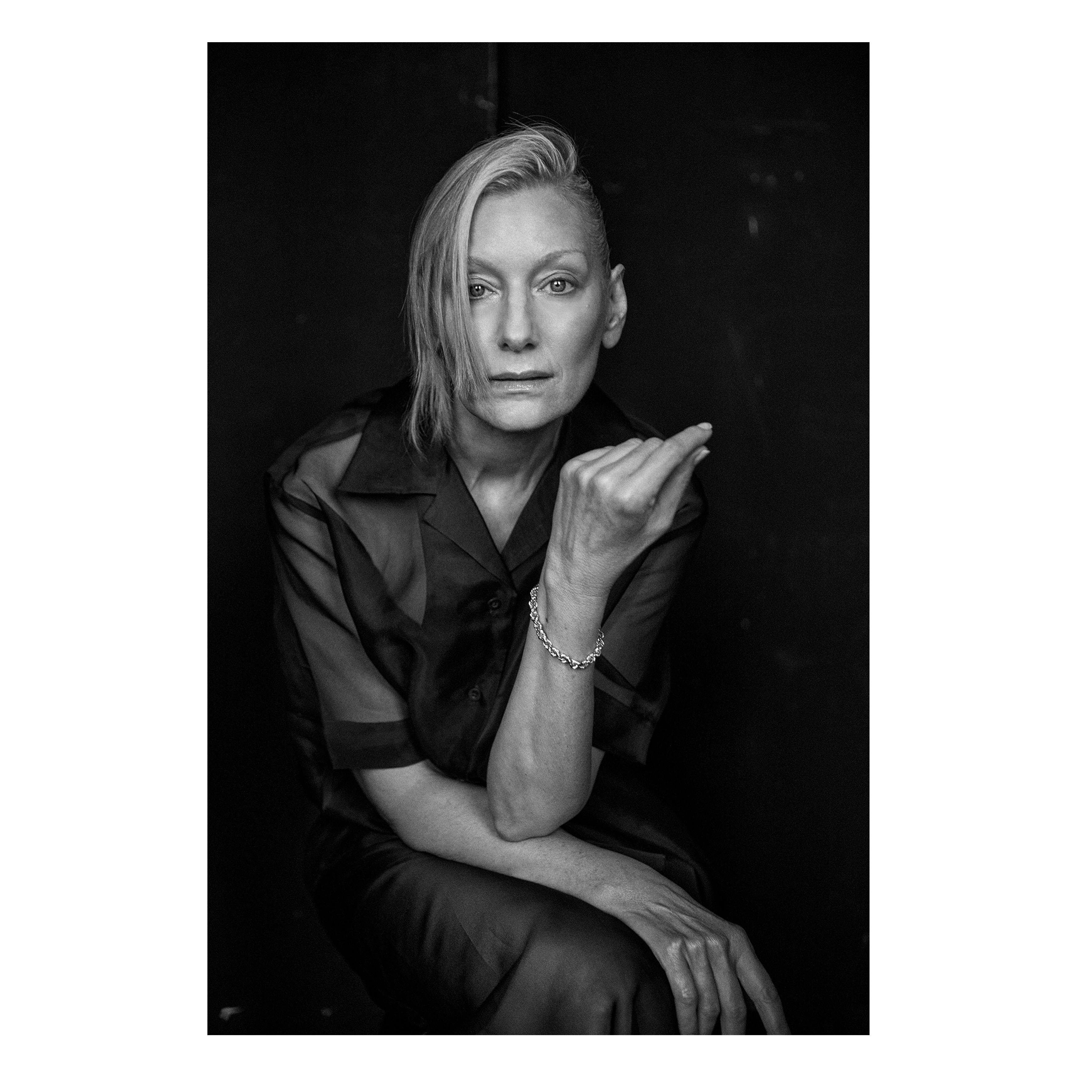 Black and white portrait of an adult female with platinum blonde hair, wearing a semi-sheer black button-down shirt, seated against a dark backdrop.