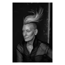 Portrait of a mature woman with windswept blonde hair wearing a sheer black top against a dark background