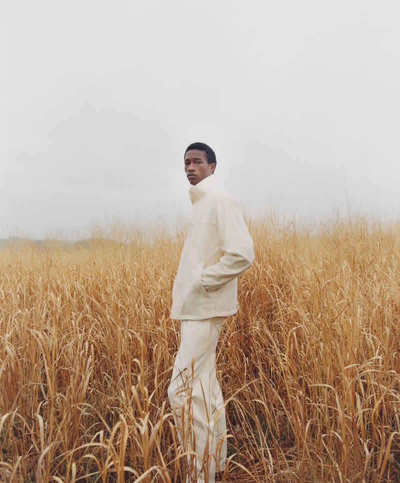 Man in cream-colored outfit stands in golden field of tall grass under an overcast sky, looking to the side with a neutral expression.