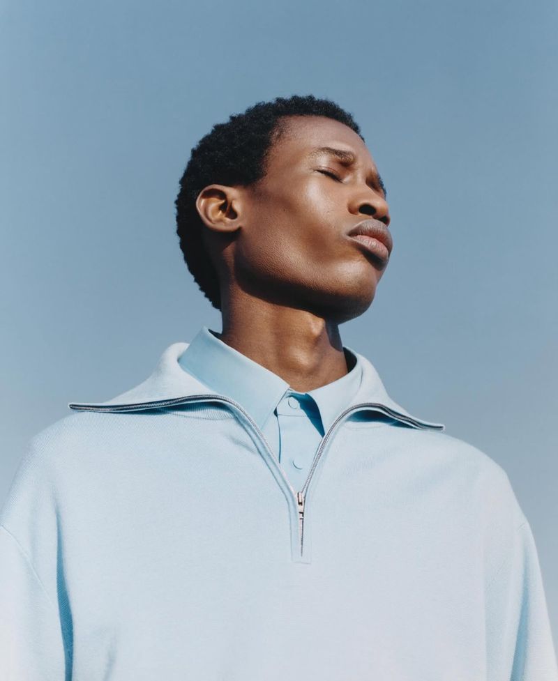 An adult man with dark hair stands outdoors against a blue sky, eyes closed, wearing a blue sweater and collared shirt. Fashion shot.