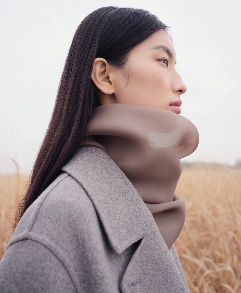 Woman in gray coat and brown scarf in outdoor setting featuring field of wheat; close-up of a young woman's profile, fashion editorial.