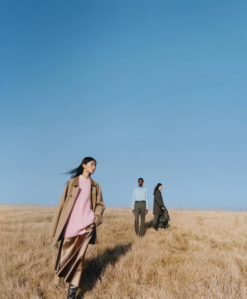 Three fashion models in coats and skirts walking through a field of golden grass under a clear blue sky on a sunny day.