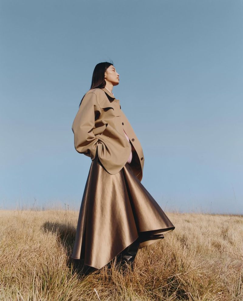 Asian woman in a tan coat and skirt posing in a field against a blue sky. Fashion shot with natural light, outdoor setting.