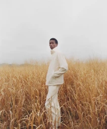 Model Daniel Awaridhe in a cream zip jacket and trousers standing in a field of tall dry grass against an overcast sky.