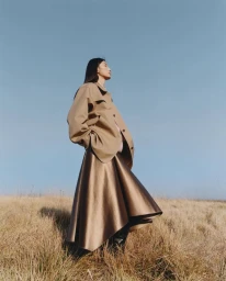 Model standing outdoors in a dry field wearing a camel jacket and flared skirt against a clear sky