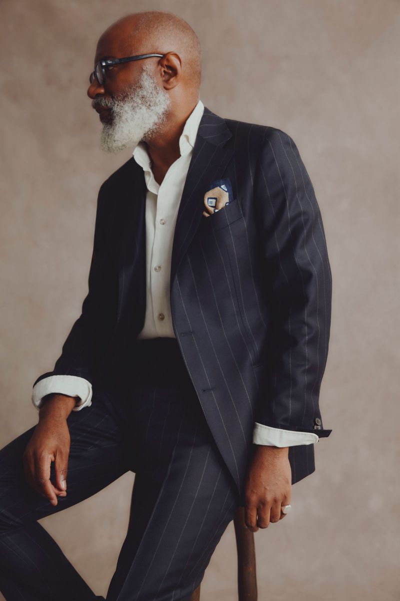 Man in a pinstripe suit with a beard posing in a studio, sitting on a stool, looking to the side, formal wear, studio lighting.
