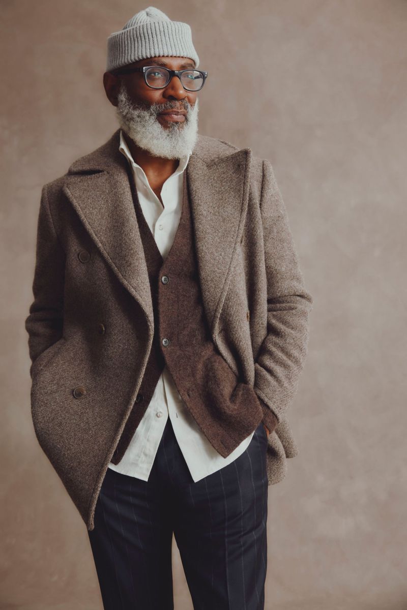 An elderly Black man with a white beard poses in a wool coat, vest, shirt, trousers, glasses, and a beanie against a neutral backdrop.
