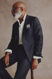 Jason Jules seated in a navy pinstripe suit and white shirt against a neutral textured studio backdrop.
