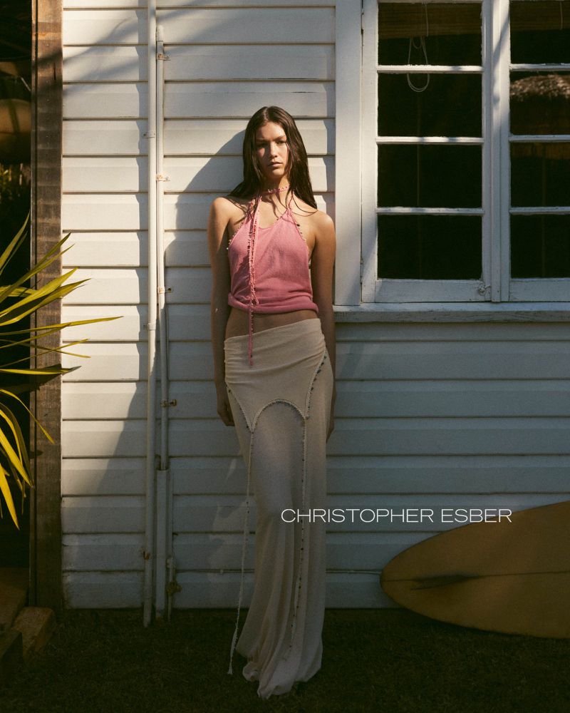 A woman stands by a white wall in a pink halter top and long skirt, featuring a surfboard and natural light.