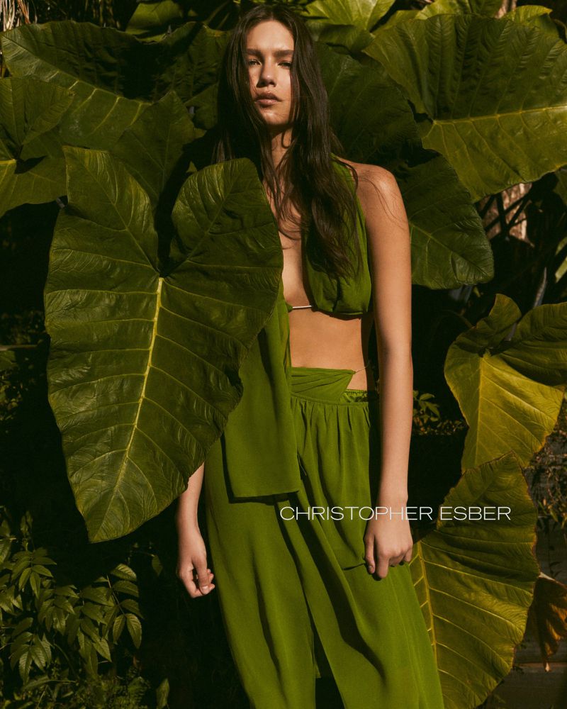Fashion editorial photo of a woman in green draped clothing surrounded by large tropical leaves, captured in natural light.