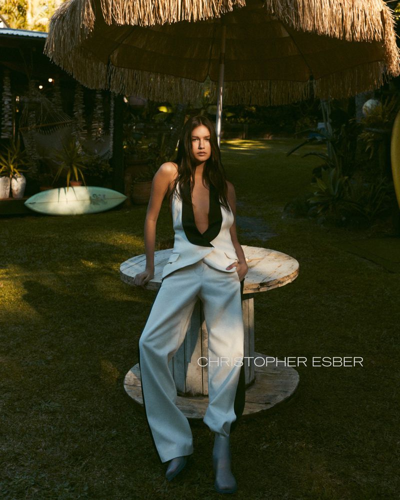 Brunette woman in a white pantsuit poses next to a vintage cable spool table topped with a fringed umbrella in an outdoor setting.