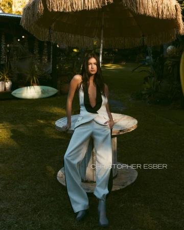 Brunette woman in a white pantsuit poses next to a vintage cable spool table topped with a fringed umbrella in an outdoor setting.