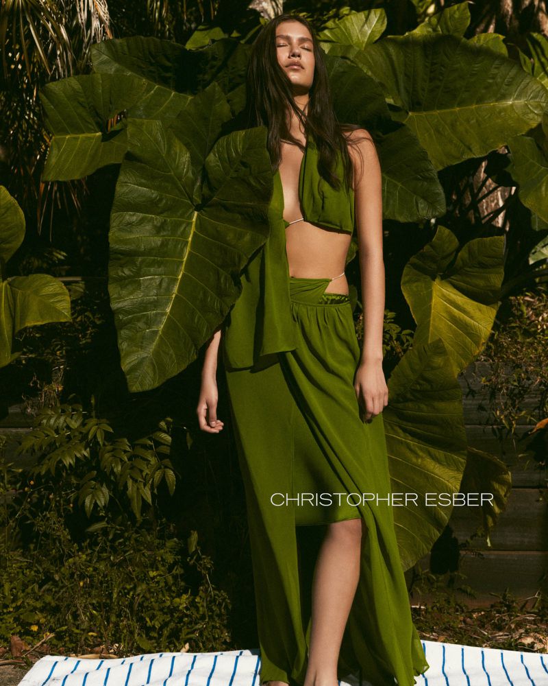 A woman poses in a green dress with large green leaves in the background, showcasing a fashion-forward and nature-inspired aesthetic.