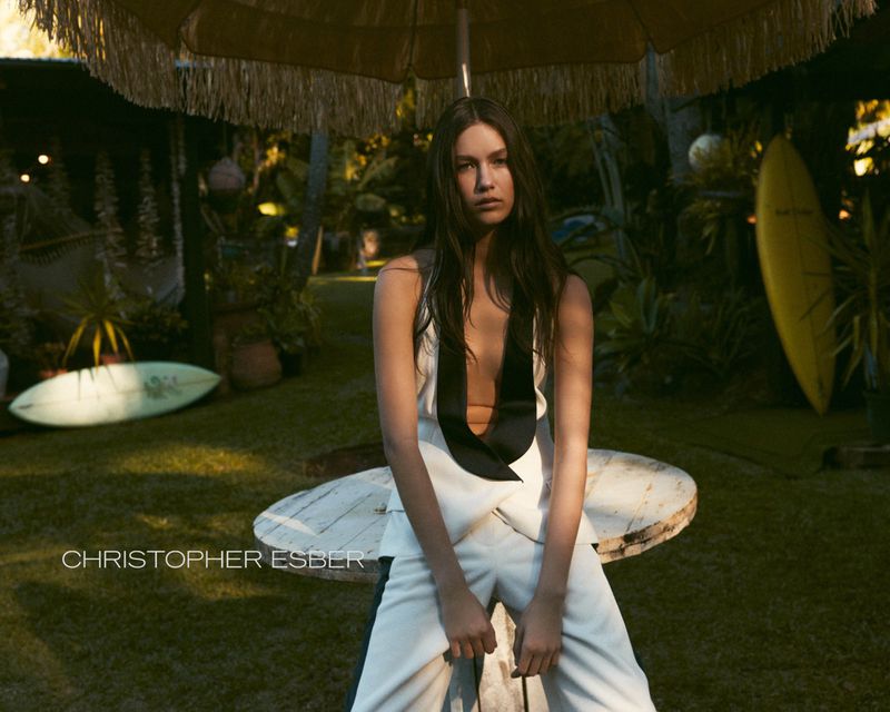 A woman in a white jumpsuit sits on a white table under a fringed umbrella, surrounded by green plants and surfboards in natural light.