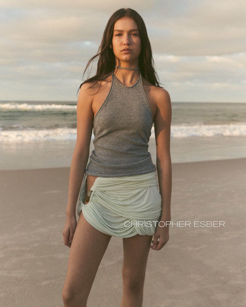 A young woman in a gray tank top and pale green skirt stands on a beach with ocean waves and a cloudy sky at dusk or dawn.