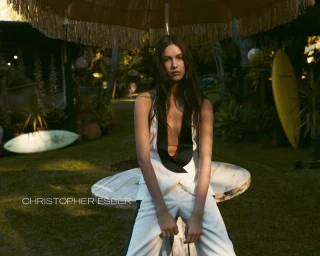 Model seated outdoors under a fringed umbrella wearing a white halter top and trousers by Christopher Esber