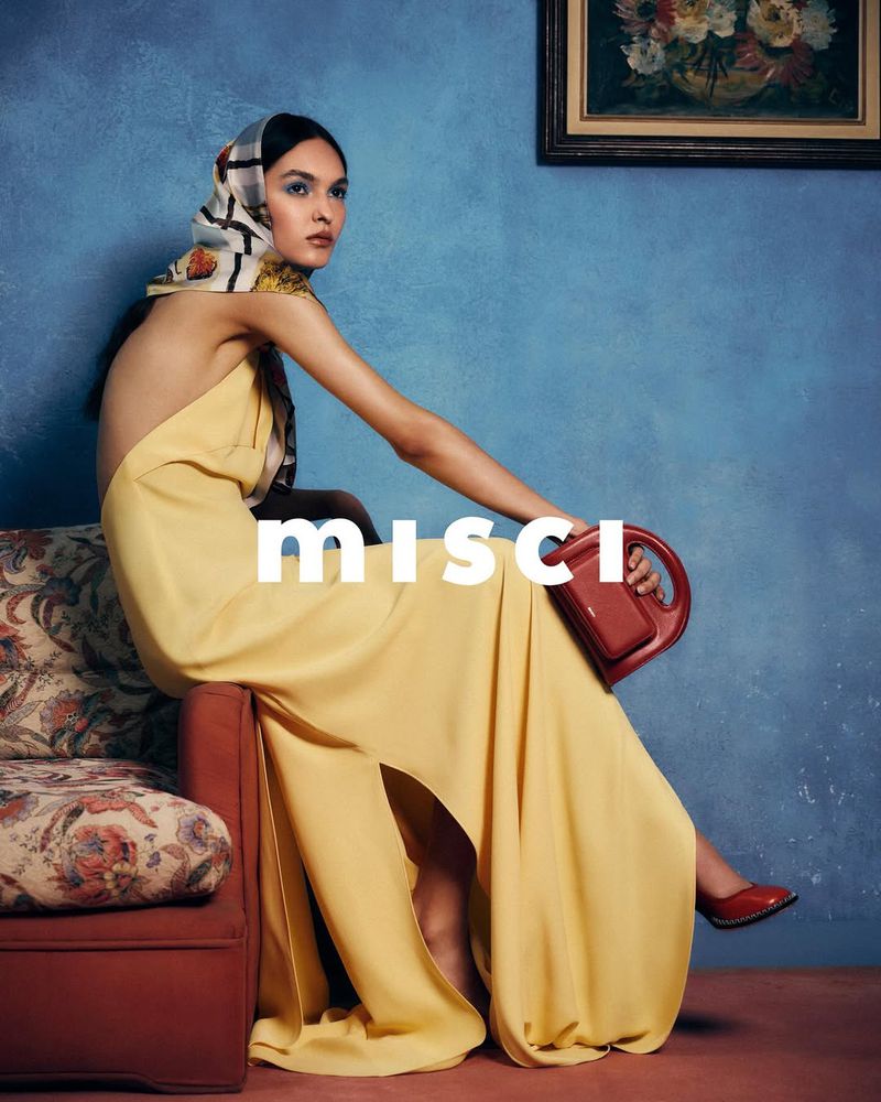 Fashion shot of a woman in a yellow dress and patterned headscarf sitting on a red sofa against a blue wall with a painting.