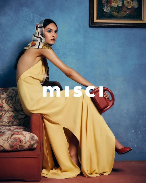 Fashion shot of a woman in a yellow dress and patterned headscarf sitting on a red sofa against a blue wall with a painting.
