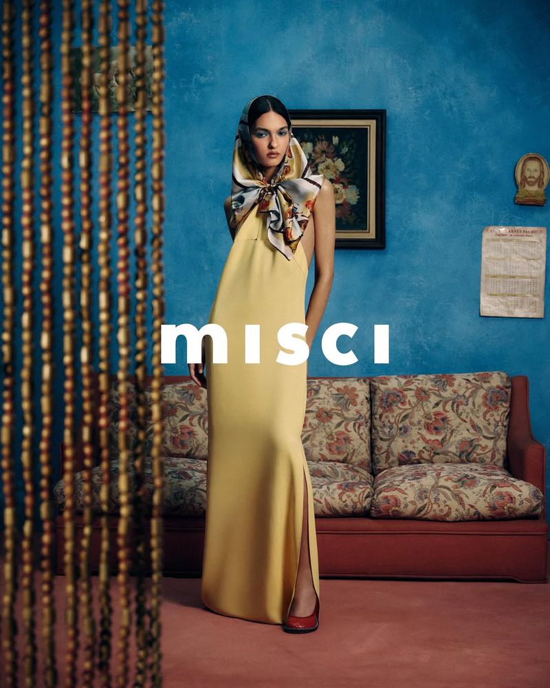 Woman in a yellow gown and headscarf stands in front of blue wall with beaded curtain, couch, and artwork in an indoor setting.