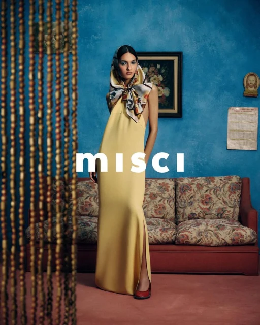 Woman in a yellow gown and headscarf stands in front of blue wall with beaded curtain, couch, and artwork in an indoor setting.