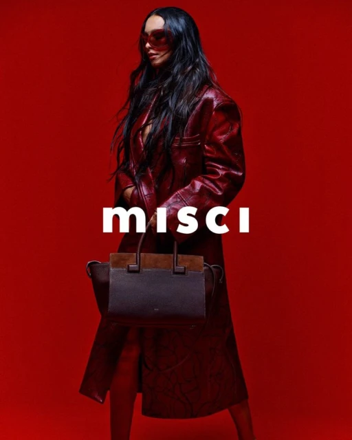 Fashionable adult woman in a red coat and sunglasses poses in front of a red background holding a dark brown handbag.