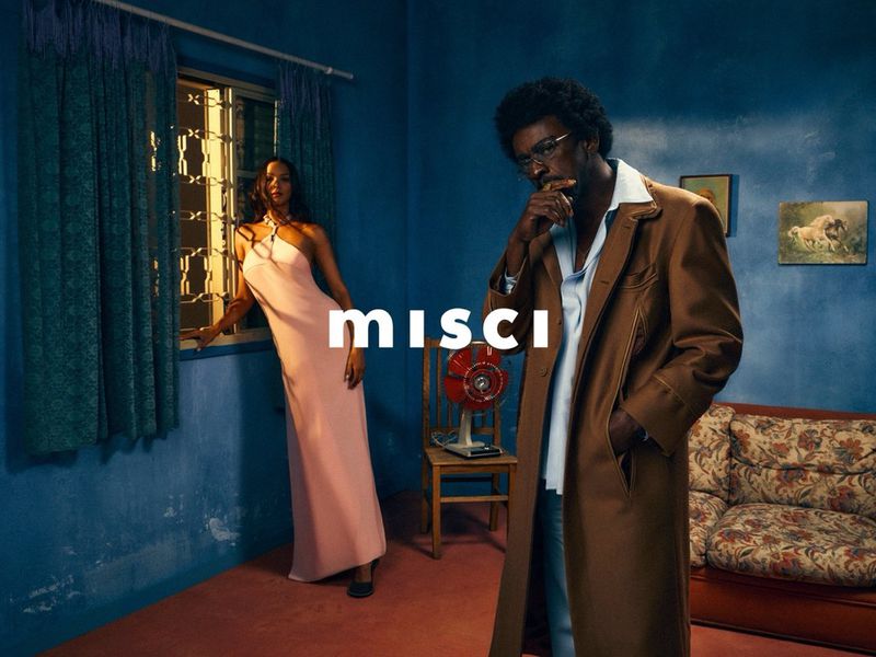 Fashion shot of a man in a trench coat and a woman in a gown, posing indoors in a room with vintage decor and blue walls.