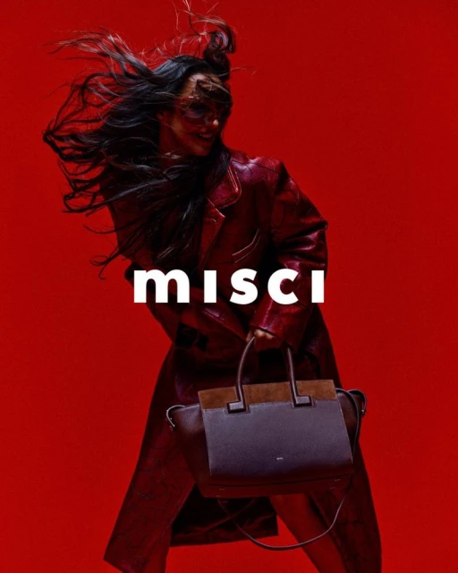 A woman in a red leather trench coat poses in front of a red backdrop while holding a dark handbag, fashion-forward and chic.