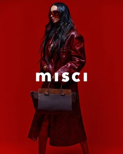 Model with long dark hair wearing a red leather coat and sunglasses holding a dark handbag against a solid red backdrop