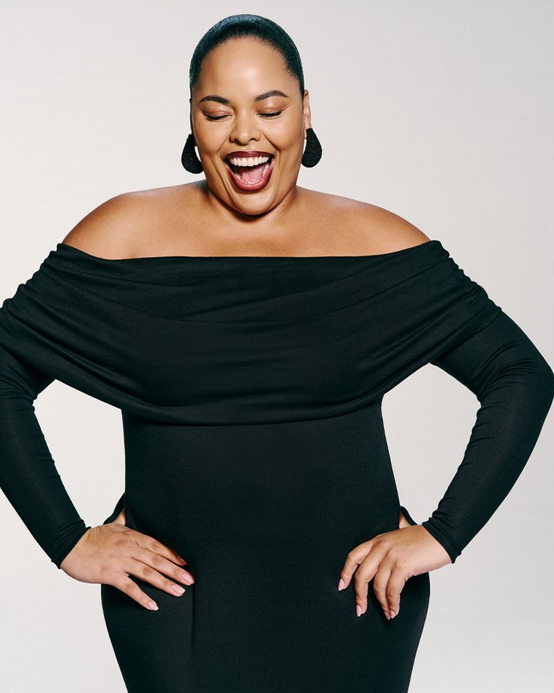 Adult woman smiles broadly in a black, off-the-shoulder, long-sleeve dress, with her hands on her waist in a photo studio setting.