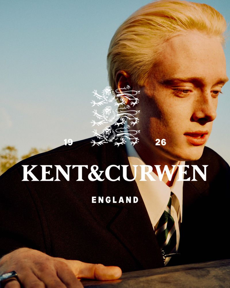 Close-up of a fair-skinned adult male model wearing a suit and tie for a Kent & Curwen fashion advertisement against a blue sky.