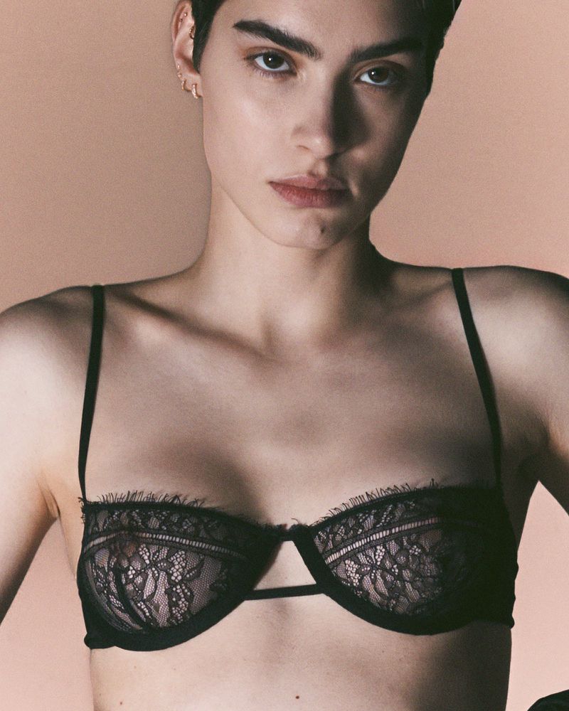 Headshot of a female model in a black lace bra against a tan background, showcasing lingerie in a studio fashion setting.