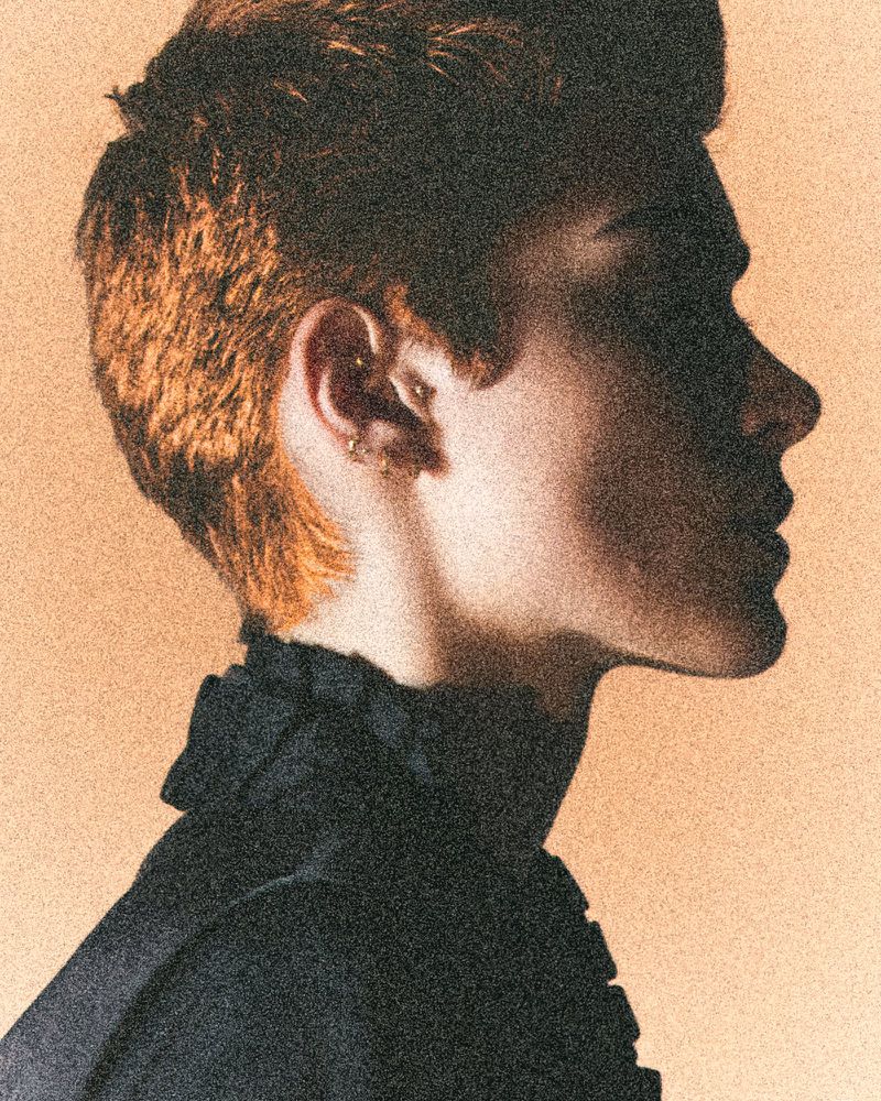 Profile portrait of a red-haired adult male in black ruffled shirt against a light peach backdrop. The lighting is dramatic.