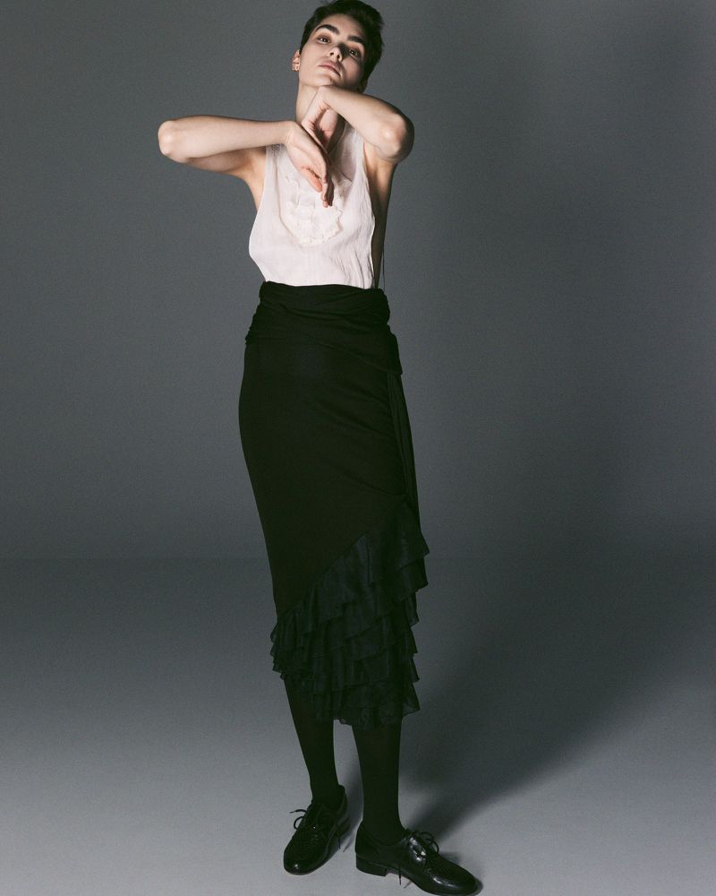 Model Snezana Gasic posing in a white sleeveless top and tiered black maxi skirt with oxford shoes against a grey studio backdrop.