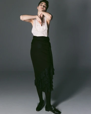 Model Snezana Gasic posing in a white sleeveless top and tiered black maxi skirt with oxford shoes against a grey studio backdrop.