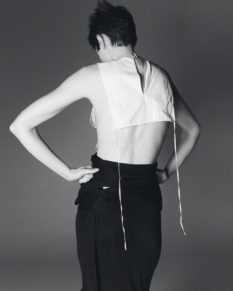 Back view of a slender woman with short hair wearing a white top and black skirt, posing against a grey studio backdrop. 
