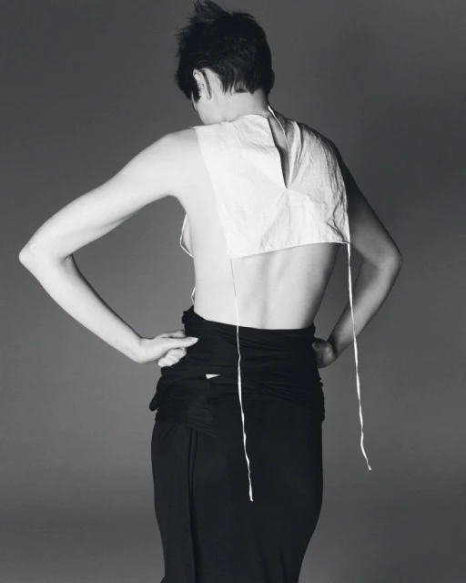 Back view of a slender woman with short hair wearing a white top and black skirt, posing against a grey studio backdrop. 