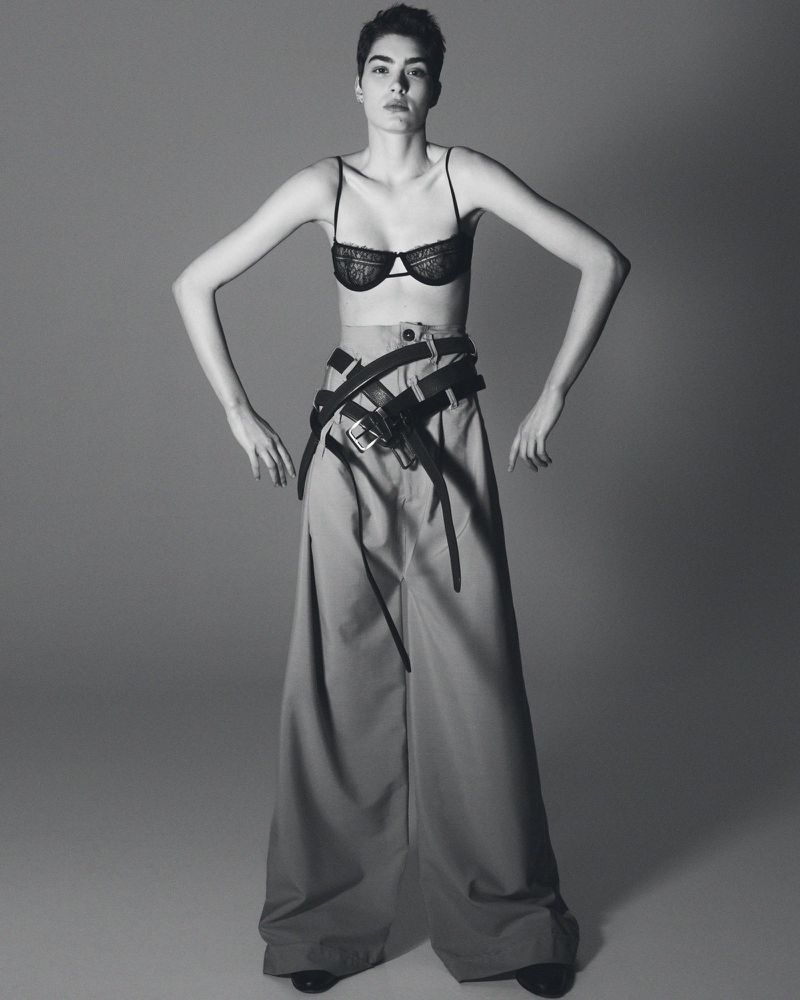 A stylish woman in a bra and wide-legged pants poses confidently in a studio shot. She wears multiple belts, emphasizing a modern, edgy fashion statement.