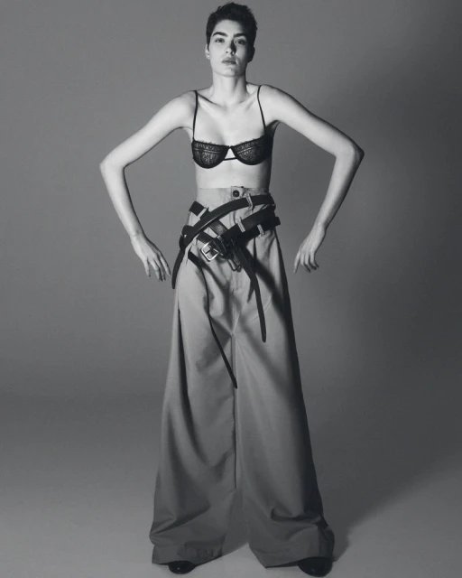 A stylish woman in a bra and wide-legged pants poses confidently in a studio shot. She wears multiple belts, emphasizing a modern, edgy fashion statement.