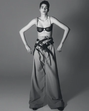 A stylish woman in a bra and wide-legged pants poses confidently in a studio shot. She wears multiple belts, emphasizing a modern, edgy fashion statement.