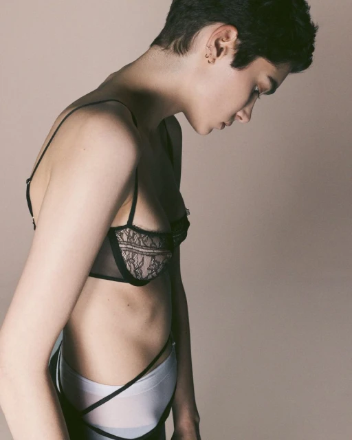 Young adult female with short hair poses in black lingerie and white shorts against a neutral backdrop in a fashion-style photo.
