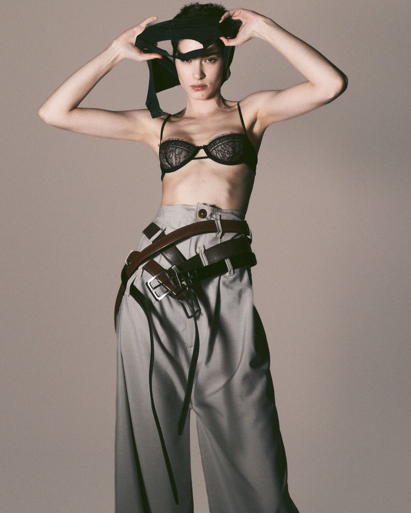 Portrait of a woman in a bra and wide-legged pants, holding a black garment over her head in a photo studio against a neutral background.