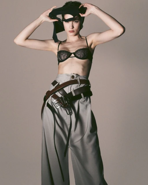 Portrait of a woman in a bra and wide-legged pants, holding a black garment over her head in a photo studio against a neutral background.