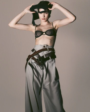 Portrait of a woman in a bra and wide-legged pants, holding a black garment over her head in a photo studio against a neutral background.