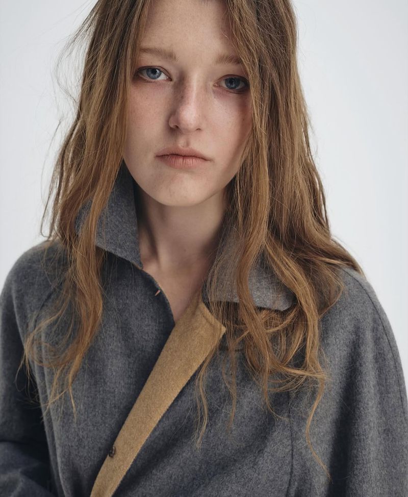 Close-up of a young woman with long brown hair wearing a gray coat with a tan strap, against a plain white background.
