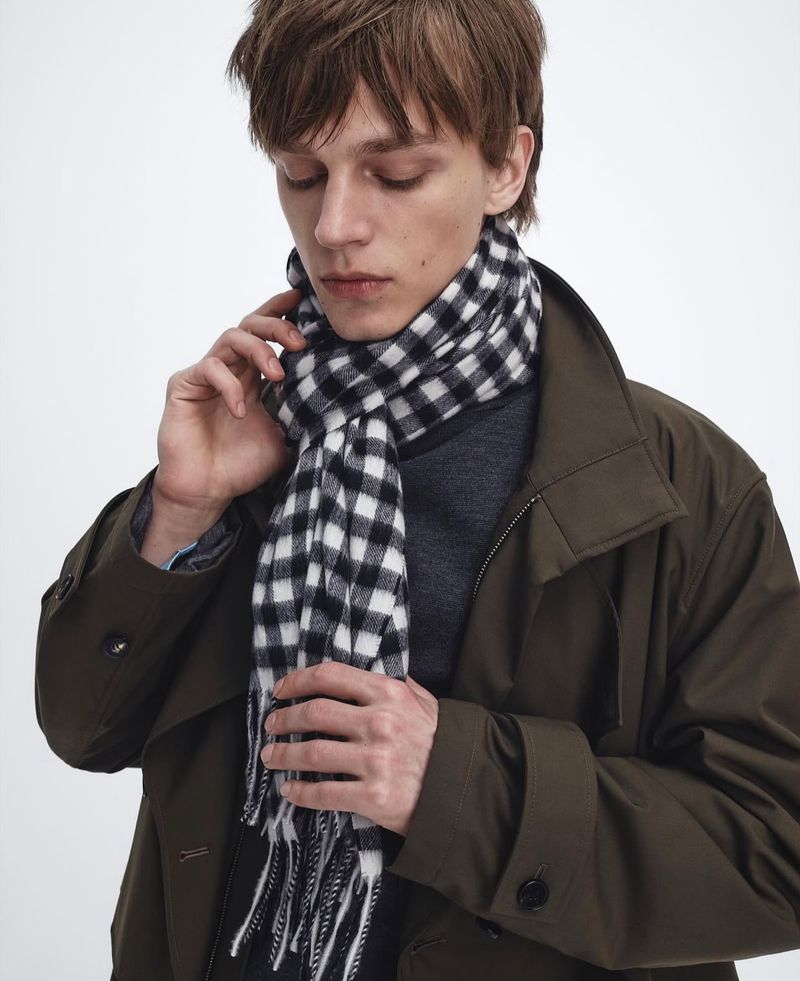 Portrait of a fair-skinned young adult male wearing a black-and-white checkered scarf and green jacket in a brightly lit indoor setting.