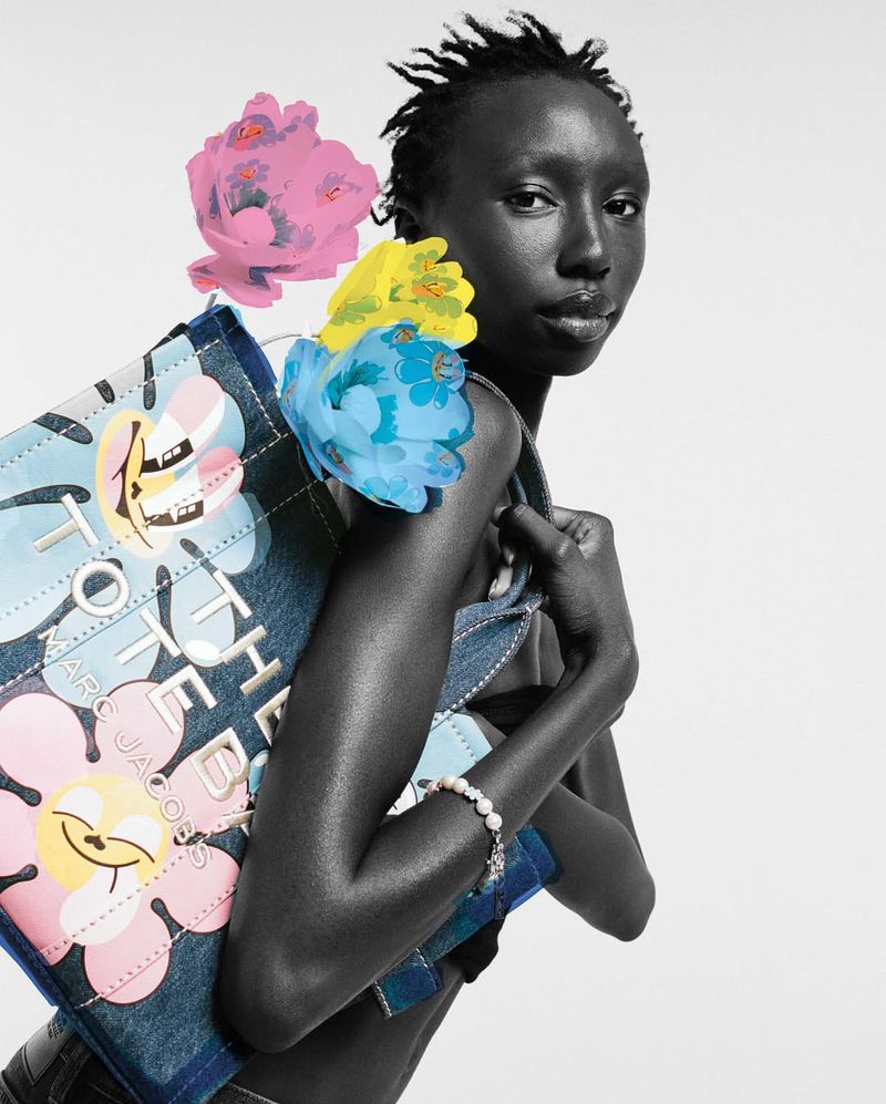 Model poses with a denim Marc Jacobs tote bag adorned with colorful floral accents against a clean, neutral background in a studio setting.