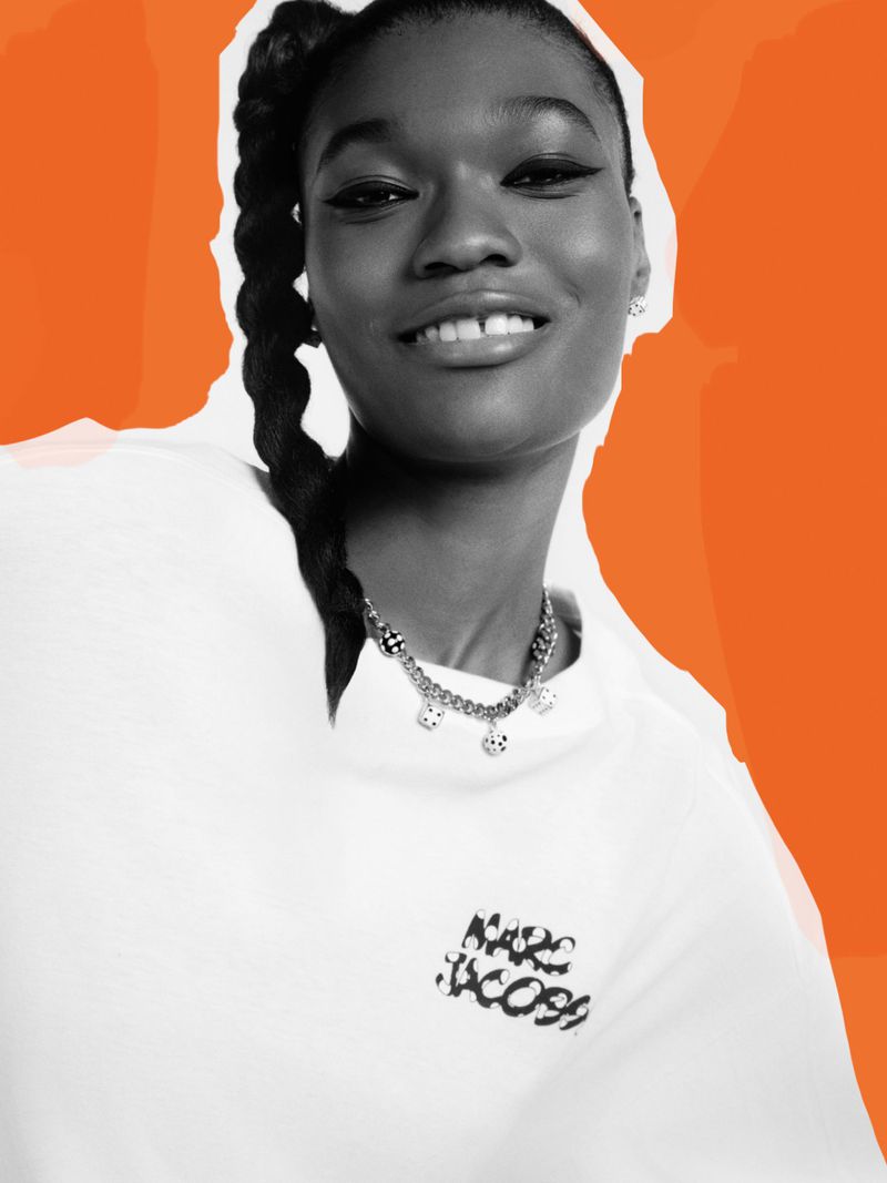 A smiling African American woman in a white top with a Marc Jacobs logo, captured in a medium shot against an orange abstract background.