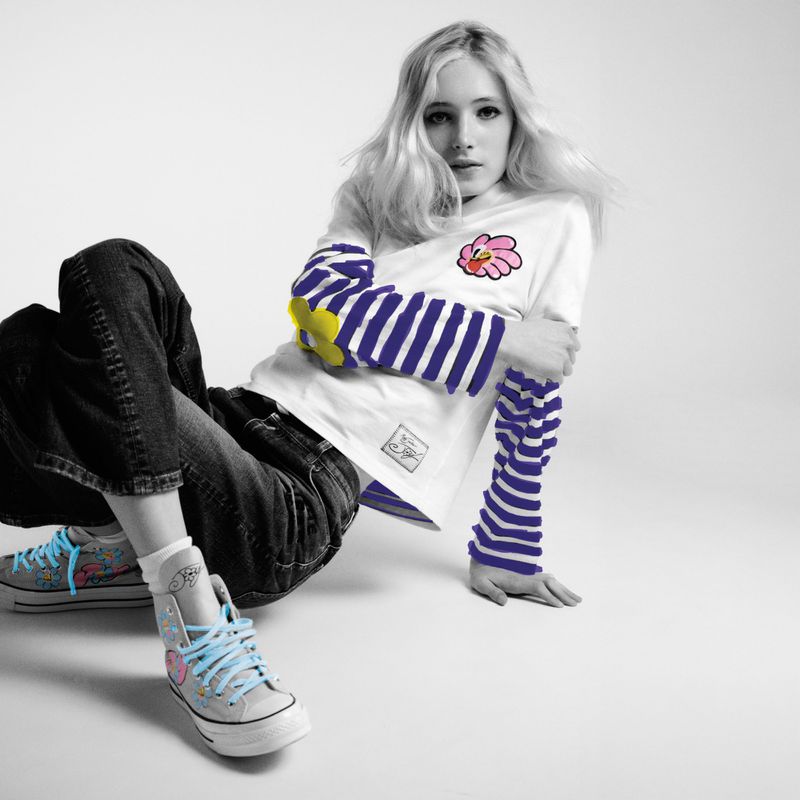 Young woman in jeans and sneakers poses in a photo studio. She wears a white t-shirt, striped sleeves, and colorful sneakers.